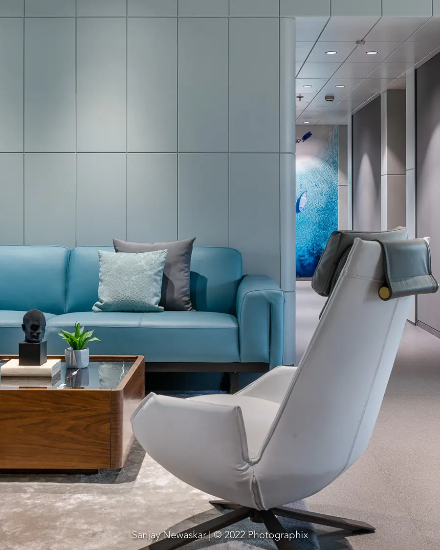 The Cadbury House: a pale blue sofa and a white lounge chair under a gridded ceiling, with an abstract blue artwork beyond.