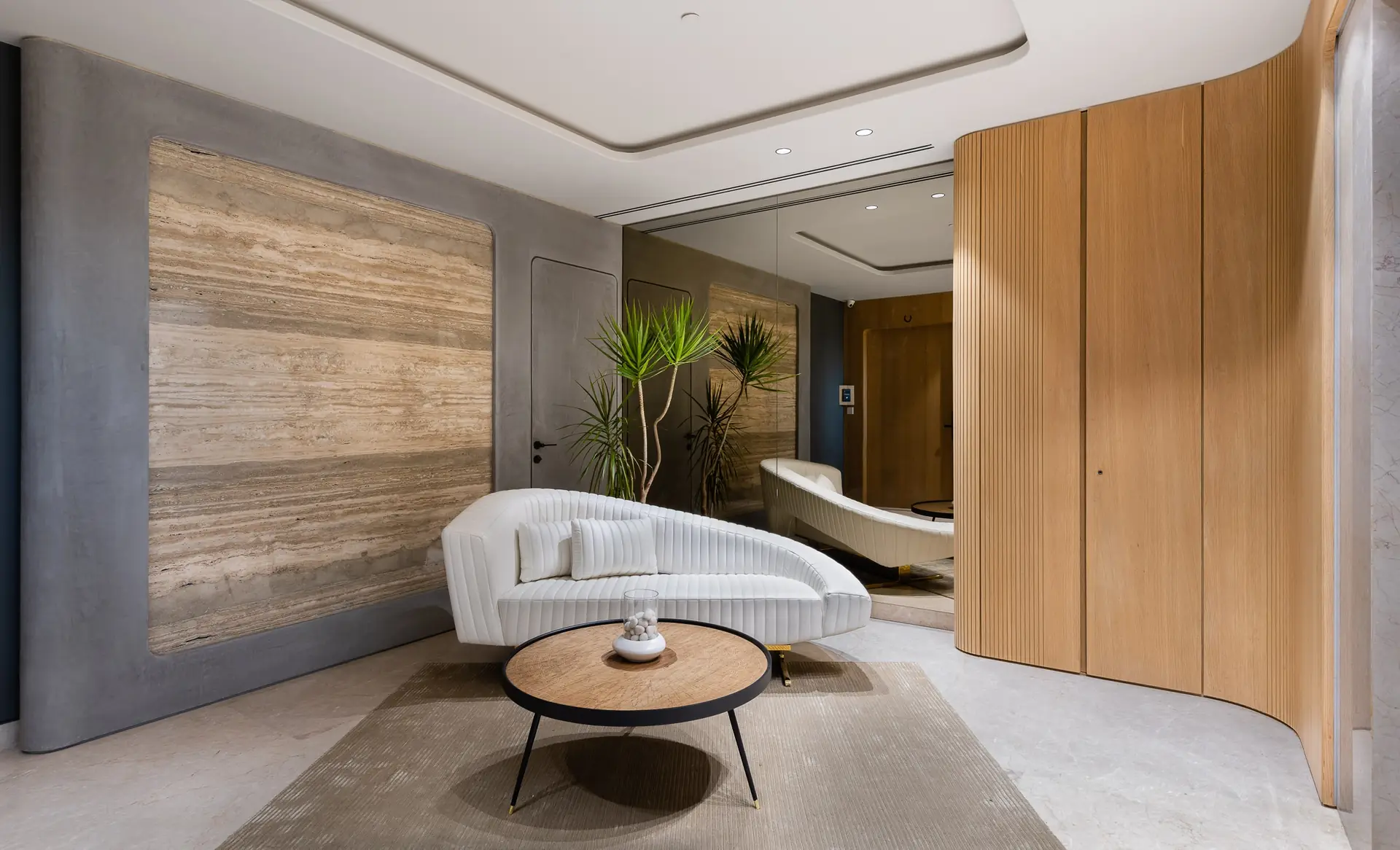 Round Edges House, Pune: a curved white sofa in front of a travertine wall, with fluted timber panelling and a round timber table.